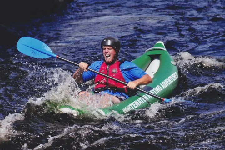 Person kayaking in whitewater rapids, wearing a helmet and life vest, paddling energetically.