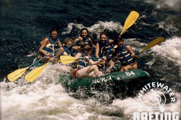 Group rafting on turbulent water, wearing life vests, smiling, with yellow paddles.